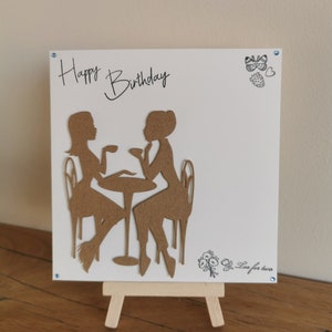 May include: A white birthday card with the words "Happy Birthday" in cursive. The card features brown silhouettes of two people at a table, with the text "Tea for two" and small illustrations. The card is displayed on a small wooden easel.