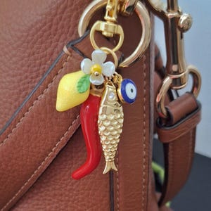 May include: A tan leather handbag with gold-tone hardware. A charm with a lemon, chilli pepper, fish, and evil eye hangs from the chain. A red gerbera daisy is placed near the bag.