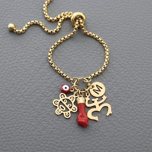 Puede incluir: Anillo ajustable dorado con diseño de cadena. El anillo presenta dijes que incluyen un ojo turco, un cuerno de coral rojo, una figura estilizada y un dije con la letra "M". El anillo se muestra sobre una superficie gris.