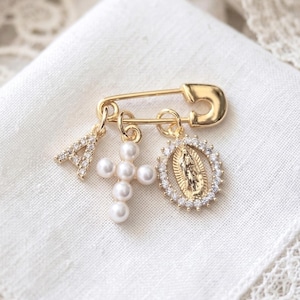 Pode incluir: Um alfinete de segurança dourado com três pingentes: uma letra "A" cravejada de strass, uma cruz de pérolas e um pingente dourado da Virgem Maria rodeado por strass. O alfinete é exibido em uma superfície branca e texturizada.