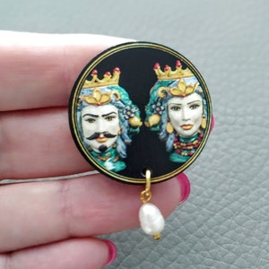 May include: A pair of earrings with a black and gold round pendant featuring a painted image of a king and queen with crowns. The pendant is attached to a gold chain with a white pearl drop.