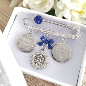 May include: A silver safety pin with a blue rhinestone charm and three dangling charms. The charms are silver and feature Arabic calligraphy and a stylized image of the Islamic symbol of faith. The pin is displayed in a white box.