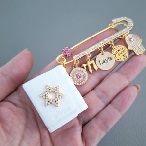 May include: Gold-colored safety pin brooch with multiple charms, including a Hamsa hand, a Star of David, and a pendant with the name "Layla". The pin is adorned with clear and pink crystals. A white fabric with a Star of David is also present.