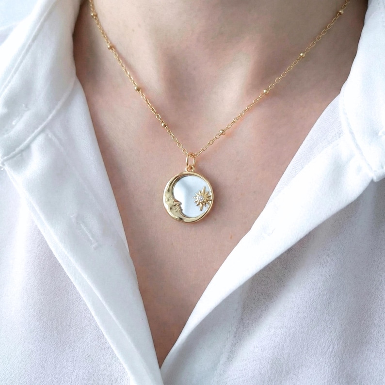 May include: A gold-toned necklace featuring a round pendant with a reflective surface. The pendant displays a crescent moon on the left side and a small, star-shaped charm with a central clear stone on the right. The chain has small gold beads.