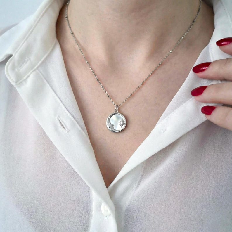 May include: A silver-toned necklace featuring a circular pendant with a relief design of a crescent moon and a sun. The pendant hangs from a delicate, thin silver-toned chain with small, evenly spaced metal beads along the length of the chain.