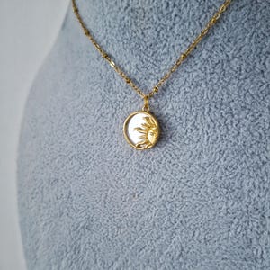 May include: A gold-toned necklace featuring a delicate chain and a circular pendant. The pendant contains a white, iridescent inlay partially covered by a raised, textured gold sun motif with rays extending across the surface of the circular charm.