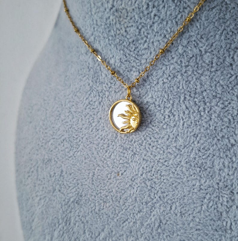 May include: A gold-toned necklace featuring a delicate chain and a circular pendant. The pendant contains a white, iridescent inlay partially covered by a raised, textured gold sun motif with rays extending across the surface of the circular charm.