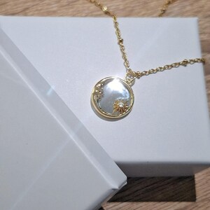 May include: A gold-toned necklace featuring a delicate chain with small beads and a circular pendant. The pendant has a reflective, pearlescent centre inlay, a gold rim, and a small gold sunburst emblem with a tiny clear stone at its centre.