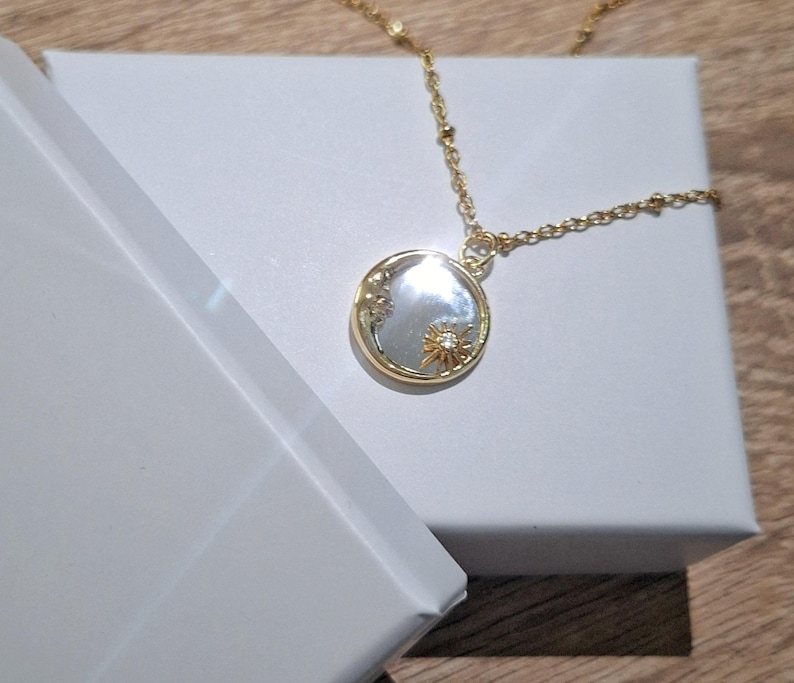 May include: A gold-toned necklace featuring a delicate chain with small beads and a circular pendant. The pendant has a reflective, pearlescent centre inlay, a gold rim, and a small gold sunburst emblem with a tiny clear stone at its centre.