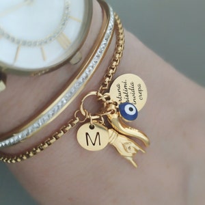 Könnte beinhalten: Goldarmband mit einem Anhänger, auf dem "fortuna, sisitimi, invidia crepa" steht, und einem blauen bösen Blick-Anhänger. Das Armband hat auch einen Anhänger mit dem Buchstaben "M" darauf.
