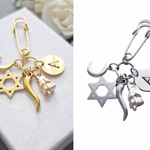 May include: Gold and silver safety pin charm with a crescent moon, star of David, chili pepper, and a hand charm. The charm also has a round disc with the letter A.