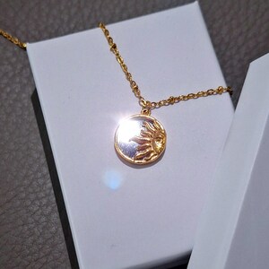 May include: A gold-toned necklace featuring a circular pendant with a raised sun motif on one side and a reflective surface on the other. The pendant hangs from a delicate gold-toned chain with small, evenly spaced beads along the length of the chain.