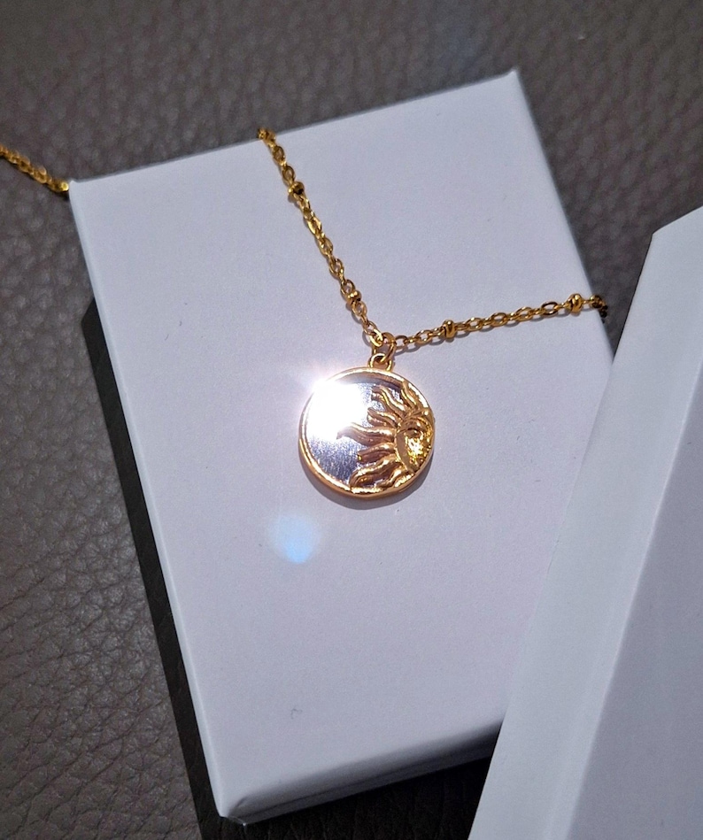 May include: A gold-toned necklace featuring a circular pendant with a raised sun motif on one side and a reflective surface on the other. The pendant hangs from a delicate gold-toned chain with small, evenly spaced beads along the length of the chain.