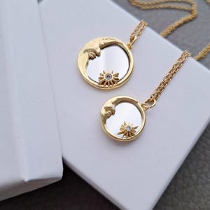 May include: Two gold-toned necklaces featuring circular pendants with a reflective mirror centre. Each pendant displays a crescent moon on one side and a small, star-shaped charm with a clear stone at the bottom, suspended on a delicate gold chain.