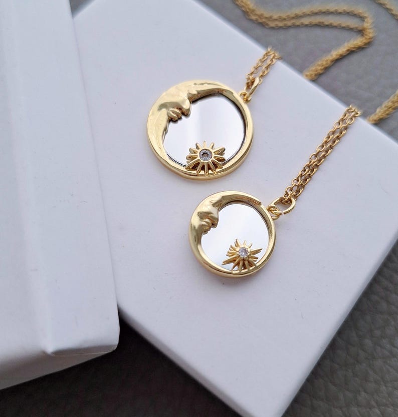 May include: Two gold-toned necklaces featuring circular pendants with a reflective mirror centre. Each pendant displays a crescent moon on one side and a small, star-shaped charm with a clear stone at the bottom, suspended on a delicate gold chain.