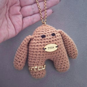 Pode incluir: Uma figura de pelúcia em crochê na cor castanho-claro pendurada numa corrente dourada. A figura apresenta dois pequenos olhos pretos, uma etiqueta oval dourada com o texto hebraico אמת e uma pulseira de corrente dourada numa perna.