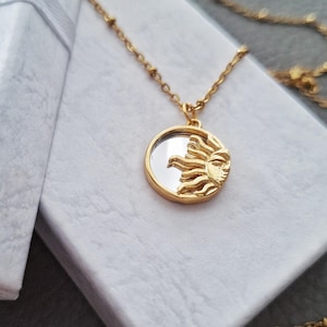 May include: A gold-toned necklace featuring a circular pendant with a reflective mirror centre and a raised, textured sun motif on one side. The pendant hangs from a delicate gold-toned chain with small, evenly spaced beads along the length.