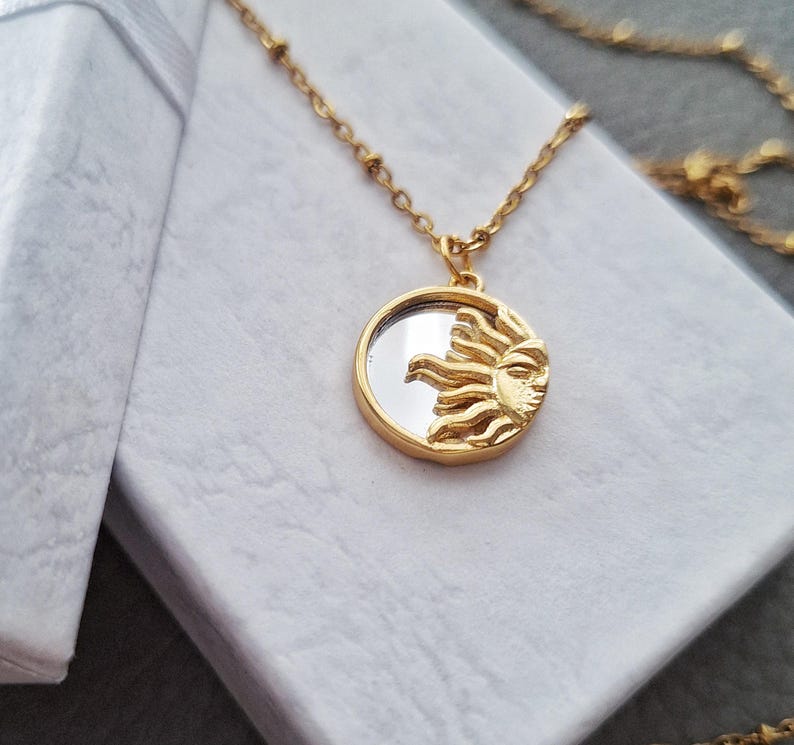 May include: A gold-toned necklace featuring a circular pendant with a reflective mirror centre and a raised, textured sun motif on one side. The pendant hangs from a delicate gold-toned chain with small, evenly spaced beads along the length.