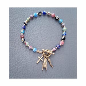 May include: A colourful beaded bracelet with glass beads and gold-coloured accents. The bracelet has a toggle clasp and three charms: a cross, a hand gesture, and a horn. The beads are in various colours including blue, pink, and black.