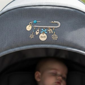 Può includere: Spilla da balia color oro con ciondoli pendenti. La spilla è adornata con cristalli trasparenti e una perla blu. I ciondoli includono una Stella di David, una mano di Hamsa, un disco con il nome "Jacob" e altri simboli religiosi. La spilla è attaccata a un passeggino grigio.