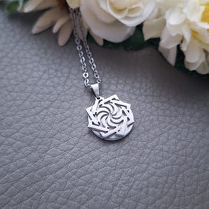 May include: A silver necklace with a geometric pendant featuring a swirling design. The pendant is made of metal and has a detailed, intricate pattern.