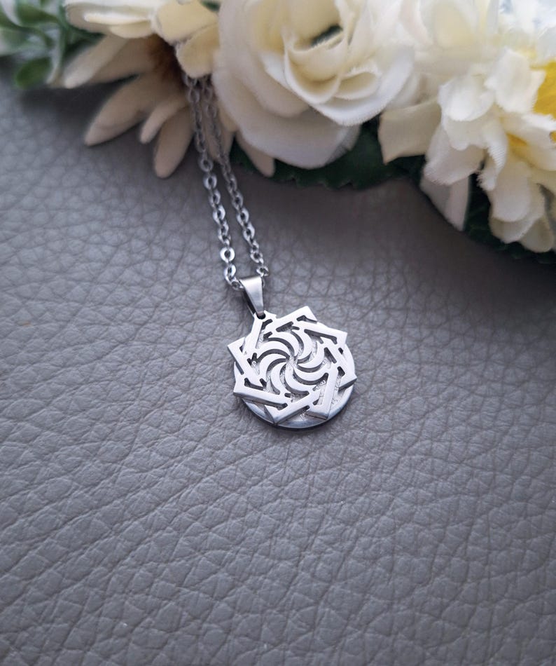 May include: A silver necklace with a geometric pendant featuring a swirling design. The pendant is made of metal and has a detailed, intricate pattern.