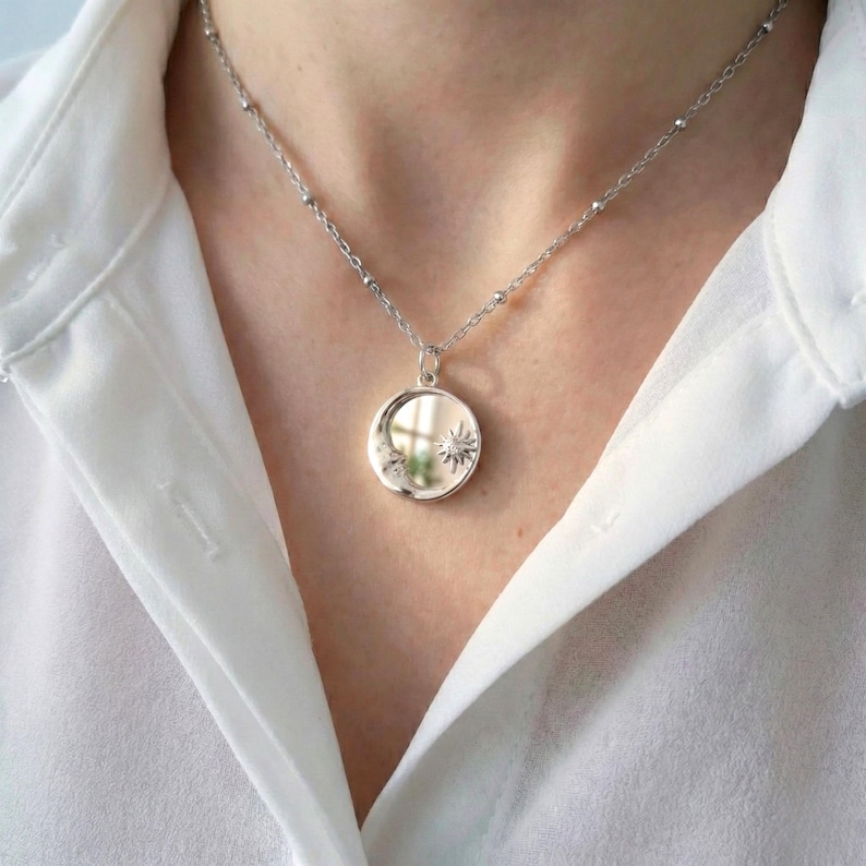 May include: A silver-coloured necklace featuring a circular pendant with a polished finish. The pendant is engraved with a small sunburst design on the right side and a crescent moon shape on the left. The chain consists of small, evenly spaced metal beads.
