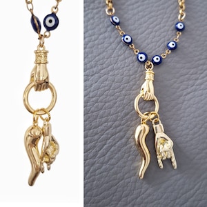 May include: Gold-coloured necklace with a chain of blue and white evil eye beads. The pendant features a hand holding a ring, with charms of a horn and a hand making a rock on gesture. The necklace is shown on a grey surface.