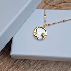 May include: A gold-toned necklace featuring a circular pendant with a mother-of-pearl inlay. The pendant displays a raised gold crescent moon on one side and a small gold sunburst with a central clear stone on the other, suspended from a delicate gold chain.