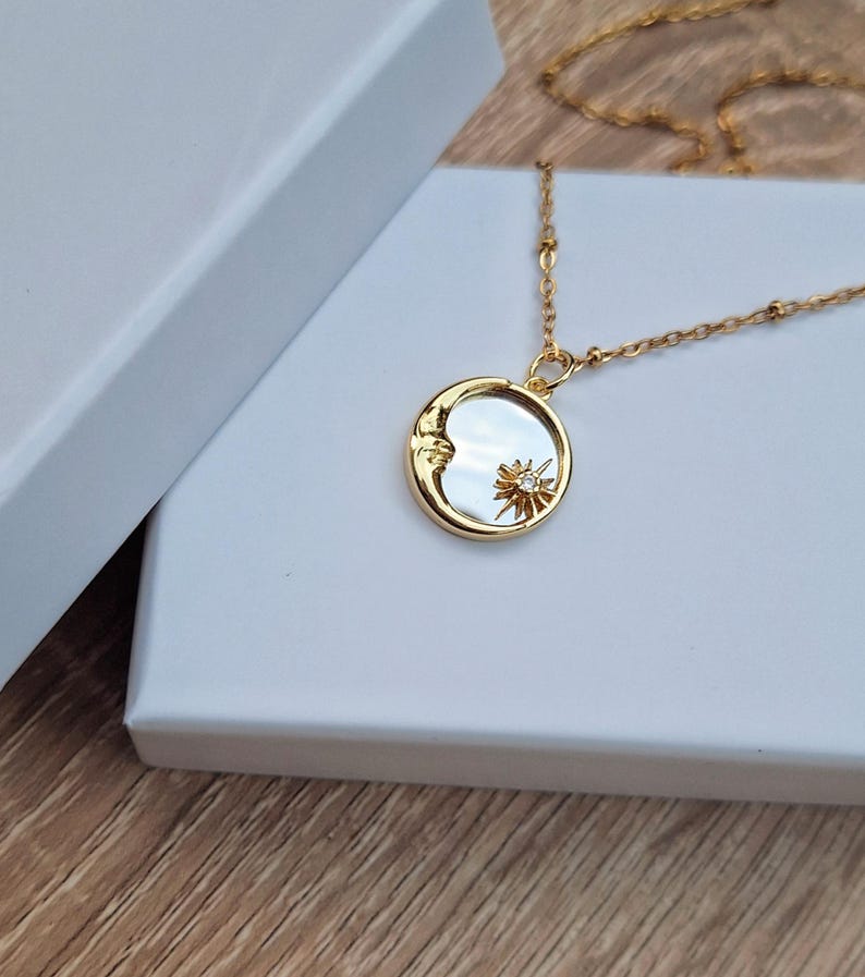 May include: A gold-toned necklace featuring a circular pendant with a mother-of-pearl inlay. The pendant displays a raised gold crescent moon on one side and a small gold sunburst with a central clear stone on the other, suspended from a delicate gold chain.