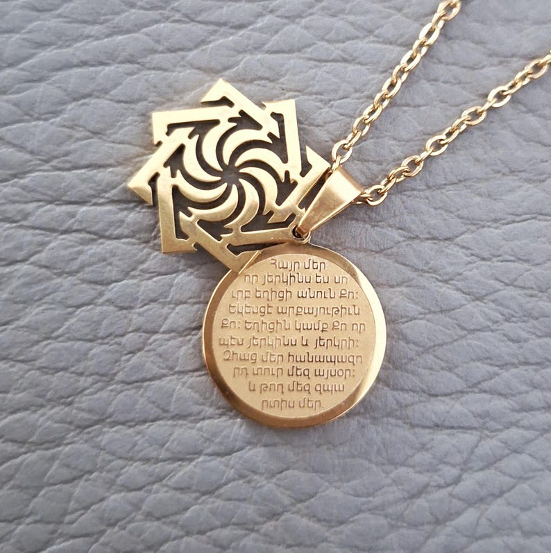 May include: Gold necklace with a pendant featuring a stylized eight-pointed star and a round charm with Armenian text. The text reads: "Հայր մեր որ յերկինս ես un ւրբ եղիցի անուն Քո։ Եկեսցէ արքայութիւն Քո։ Եղիցին կամք Քո որ պէս յերկինս և յերկրի: Զհաց մեր հանապազո րդ տուր մեզ այսօր: և թող մեզ զպ արտիս մեր"