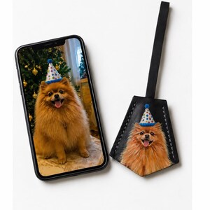 May include: A black smartphone displaying a photograph of a fluffy orange Pomeranian dog wearing a blue-trimmed party hat with colorful polka dots, next to a matching black leather luggage tag featuring the same dog image and a long strap.