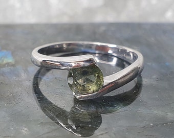 Op de afbeelding: Een zilveren ring met een groene edelsteen in een uniek, gebogen ontwerp.