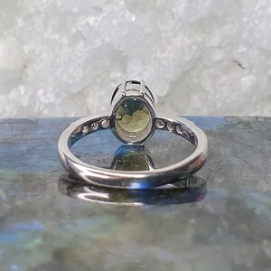 May include: A silver ring with a large oval-shaped green gemstone set in a prong setting. The ring has a band of smaller clear gemstones.