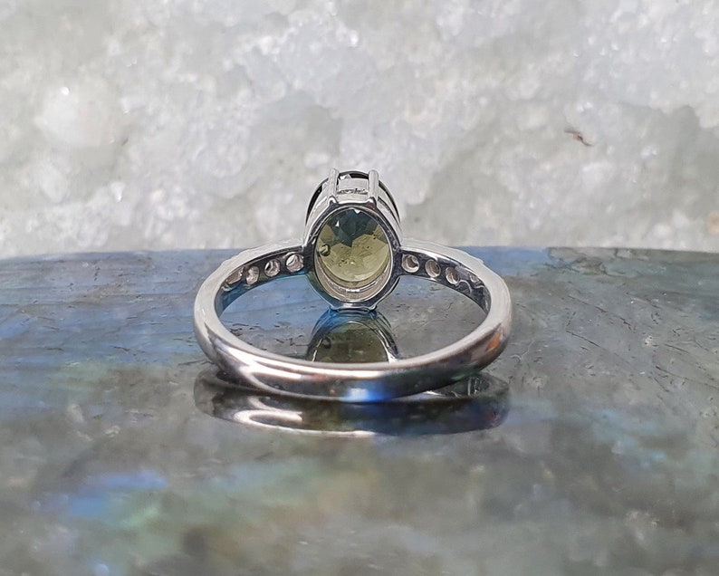 May include: A silver ring with a large oval-shaped green gemstone set in a prong setting. The ring has a band of smaller clear gemstones.