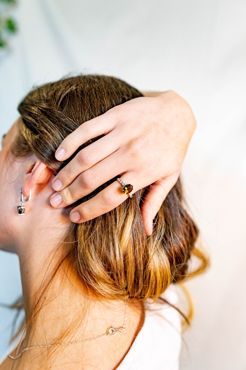 May include: A woman wearing a silver necklace and a ring with a green gemstone. The ring is on her right hand, and she is holding her hair with her left hand.