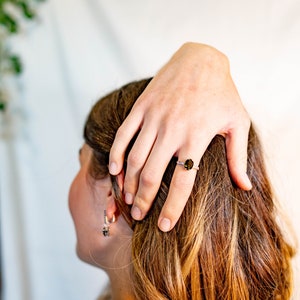 May include: A close-up of a woman's hand wearing a silver ring with a dark green oval gemstone. The ring is on the middle finger of the hand. The woman's hair is brown and she is wearing a white shirt.