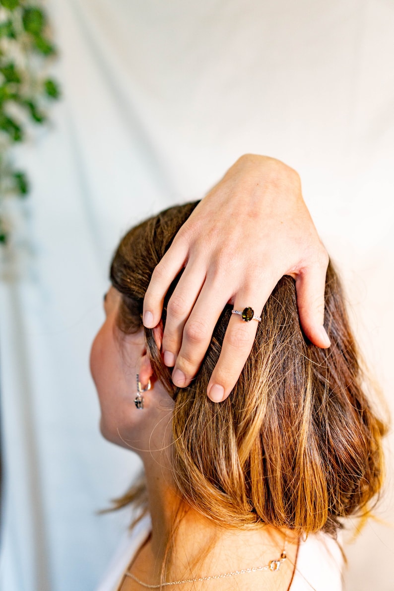 May include: A close-up of a woman's hand wearing a silver ring with a dark green oval gemstone. The ring is on the middle finger of the hand. The woman's hair is brown and she is wearing a white shirt.