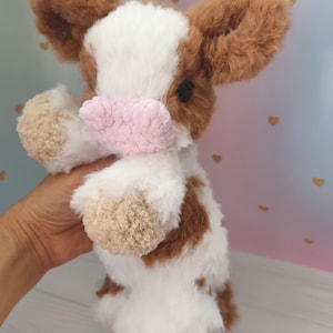 Pode incluir: Um brinquedo de vaca de pel&uacute;cia com pelo marrom e branco, um nariz rosa e orelhas marrons. O animal de pel&uacute;cia feito &agrave; m&atilde;o &eacute; segurado em frente a um fundo macio. O brinquedo &eacute; um design fofo e aconchegante.