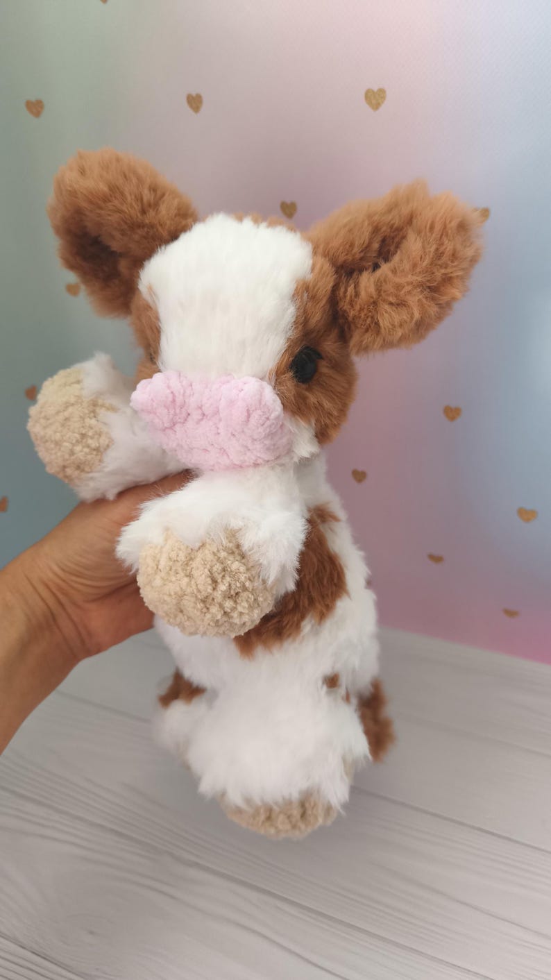 Pode incluir: Um brinquedo de vaca de pel&uacute;cia com pelo marrom e branco, um nariz rosa e orelhas marrons. O animal de pel&uacute;cia feito &agrave; m&atilde;o &eacute; segurado em frente a um fundo macio. O brinquedo &eacute; um design fofo e aconchegante.