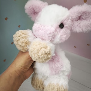 Pode incluir: Um brinquedo de vaca de pel&uacute;cia feito &agrave; m&atilde;o com uma textura macia e fofa. A vaca apresenta um corpo branco com detalhes em rosa nas orelhas, rosto e manchas. Os cascos e as patas em estilo pompom s&atilde;o castanhos. O brinquedo &eacute; segurado em uma m&atilde;o.