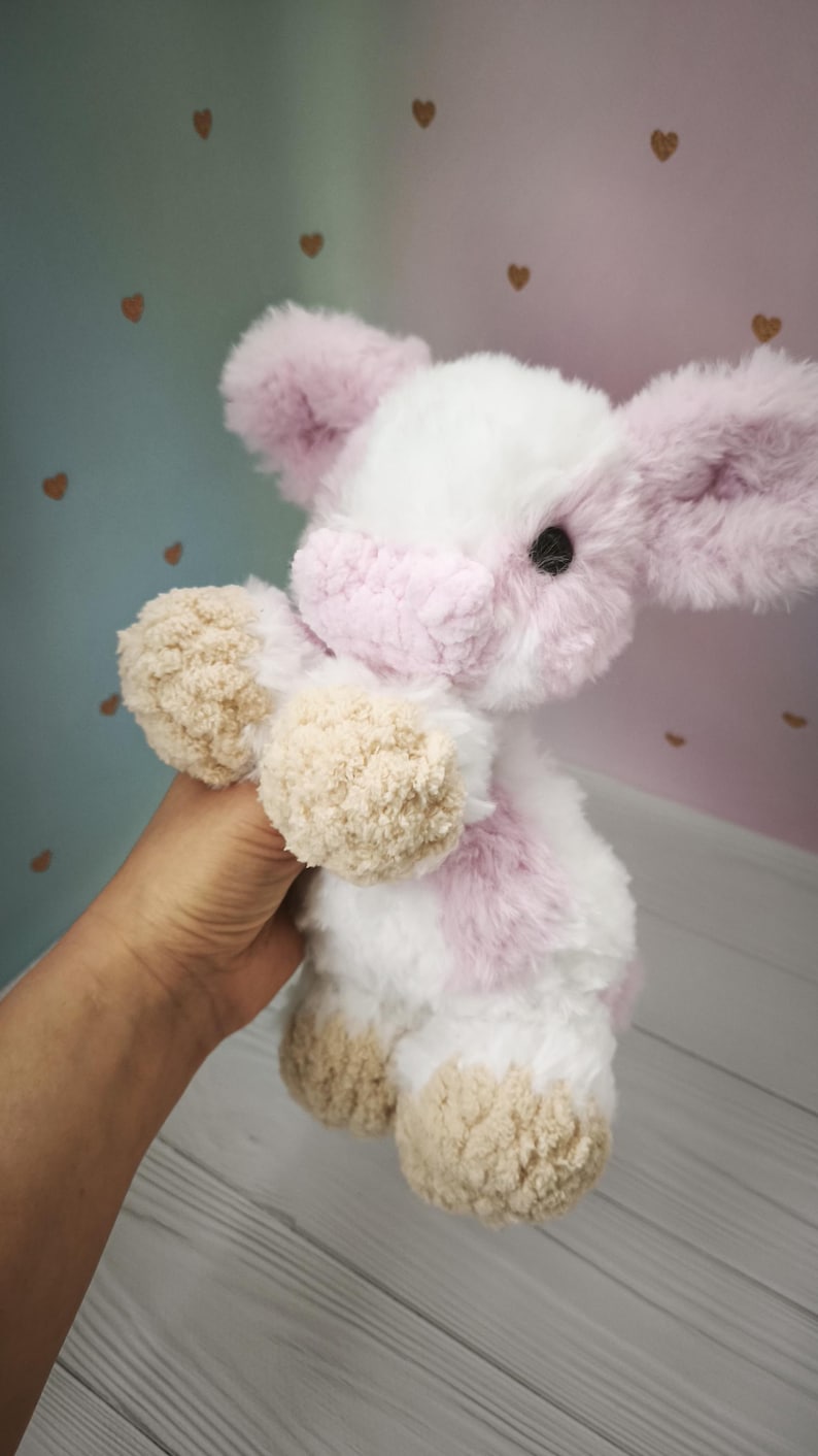 Pode incluir: Um brinquedo de vaca de pel&uacute;cia feito &agrave; m&atilde;o com uma textura macia e fofa. A vaca apresenta um corpo branco com detalhes em rosa nas orelhas, rosto e manchas. Os cascos e as patas em estilo pompom s&atilde;o castanhos. O brinquedo &eacute; segurado em uma m&atilde;o.
