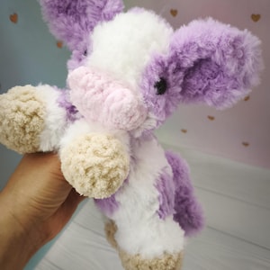 Pode incluir: Um brinquedo de pel&uacute;cia feito &agrave; m&atilde;o em forma de vaca, com um aspeto macio e texturizado. A vaca apresenta uma combina&ccedil;&atilde;o de cores branco, lavanda e bege. As orelhas e as manchas s&atilde;o lavanda, o corpo &eacute; branco e os cascos s&atilde;o bege.