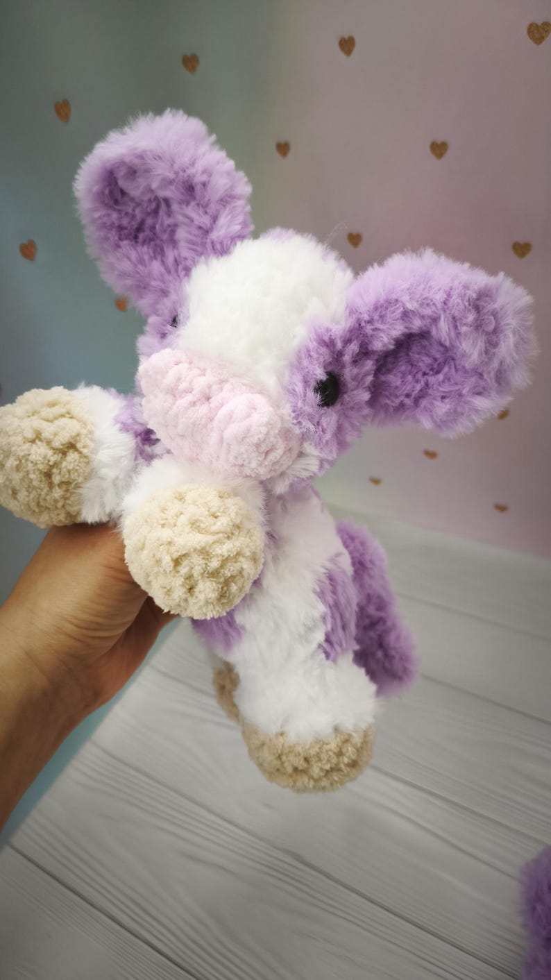 Pode incluir: Um brinquedo de pel&uacute;cia feito &agrave; m&atilde;o em forma de vaca, com um aspeto macio e texturizado. A vaca apresenta uma combina&ccedil;&atilde;o de cores branco, lavanda e bege. As orelhas e as manchas s&atilde;o lavanda, o corpo &eacute; branco e os cascos s&atilde;o bege.