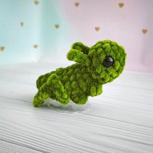 May include: A handmade, green crocheted bunny with large black eyes. The plush toy is made of a soft, textured yarn and is posed on a white surface. The background is a soft gradient of pink and blue with gold heart accents.