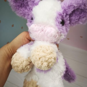 Pode incluir: Um brinquedo de pel&uacute;cia de vaca com uma textura macia e fofa. A vaca apresenta um corpo branco com orelhas e detalhes roxos e um focinho rosa. Os cascos s&atilde;o de cor bege claro. O brinquedo foi projetado para ser aconchegante e &eacute; segurado em uma m&atilde;o.