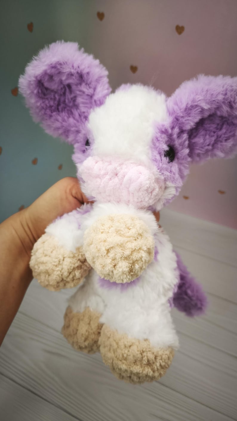 Pode incluir: Um brinquedo de pel&uacute;cia de vaca com uma textura macia e fofa. A vaca apresenta um corpo branco com orelhas e detalhes roxos e um focinho rosa. Os cascos s&atilde;o de cor bege claro. O brinquedo foi projetado para ser aconchegante e &eacute; segurado em uma m&atilde;o.
