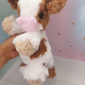 Pode incluir: Um brinquedo de pel&uacute;cia em forma de vaca com corpo branco, manchas marrons e orelhas marrons fofas. A vaca tem um nariz rosa e &eacute; segurada contra um fundo pastel com detalhes de cora&ccedil;&otilde;es dourados. O brinquedo &eacute; macio.