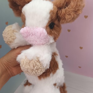 Pode incluir: Um brinquedo de vaca de pel&uacute;cia feito &agrave; m&atilde;o, com pelo castanho e branco. A vaca tem orelhas grandes e fofas castanhas, um nariz rosa e olhos pretos pequenos. Um brinquedo macio e fofinho, ideal para crian&ccedil;as ou como decora&ccedil;&atilde;o.