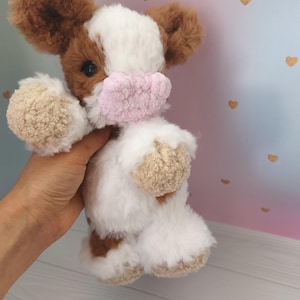 Pode incluir: Um brinquedo de pel&uacute;cia de vaca feito &agrave; m&atilde;o com pelo marrom e branco, orelhas grandes marrons e um focinho rosa. O brinquedo &eacute; segurado em uma m&atilde;o, mostrando sua textura macia e artesanato detalhado. O fundo apresenta um gradiente de rosa e azul com detalhes de cora&ccedil;&atilde;o dourado.